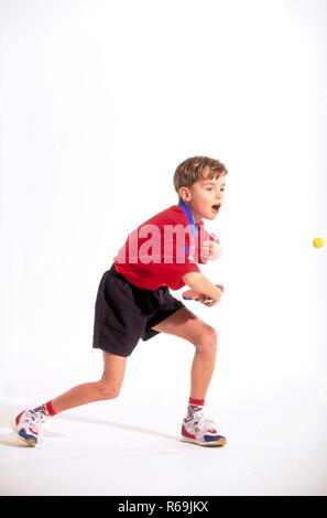 Portrait, Ganzfigur, Junge mit kurzen Haaren, 10 Jahre, in rotem Trikot und schwarzer Shorts mit Tischtennissschlaeger beim Schlagen des gelben Balles Stock Photo