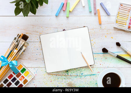 Colorful creative table with blank notebook for sketches and paints, pencil, paintbrushes set and cup of coffee on white wooden table, top view, copy  Stock Photo