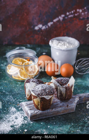 Carrot-chocolate muffin dusted with icing sugar, a cup of tea, baking ingredients. Flour, eggs, lemon citrus on a dark table Copy space Vertical Stock Photo