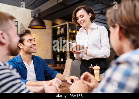 Smiling pretty waitress with short hair wearing white blouse standing at table with guests and making notes in sketchpad while taking order in restaur Stock Photo