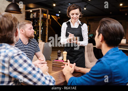 Positive smiling attractive young waitress in black apron standing at guests table and talking to male friends in loft restaurant, serious handsome me Stock Photo