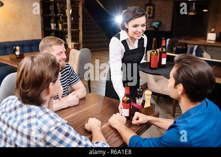 Smiling content beautiful young lady waitress in black apron giving beer to guest in pub and holding tray while talking to men, she flirting with them Stock Photo