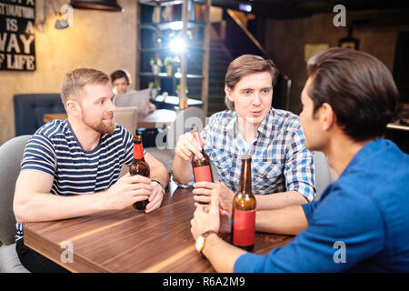 Cheerful positive young guys in casual clothing sitting at wooden table drinking beer in pub Stock Photo