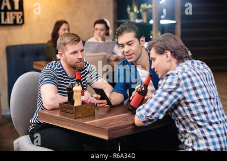 Content handsome young friends in casual clothing sitting at table in bar and drinking beer while watching video on smartphone Stock Photo