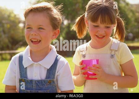 Parkszene, Portrait, Nahaufnahme, Paar, Maedchen mit kurzen Zoepfen mit pink farbenem Plastikbecher in den Haenden und Junge im Alter von 5-6 Jahren auf einer Wiese Stock Photo