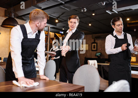 Serious displeased handsome young restaurant manager in formal jacket gesturing hand while giving task to waiter during cleanup Stock Photo