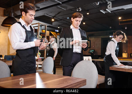 Serious concentrated handsome young restaurant manager using digital tablet while checking online records, waiting staff cleaning tables and crockery Stock Photo
