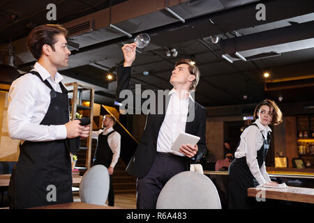 Serious professional restaurateur in black jacket holding tablet and checking cleanliness of wineglass while waiters wiping tables Stock Photo