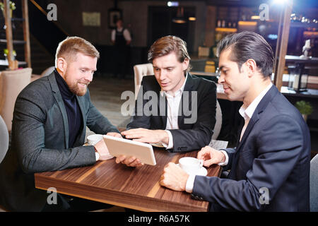 Smiling confident handsome young businessmen sitting at table and watching presentation on tablet at coffee break Stock Photo