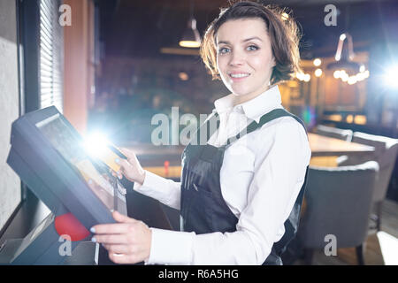 Cheerful excited beautiful young lady waitress standing in modern restaurant and using POS terminal while looking at camera Stock Photo