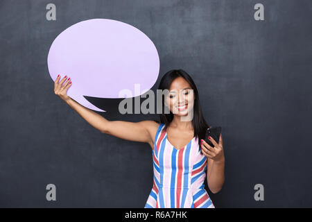 Image of charming asian woman 30s holding blank thought bubble and smartphone standing isolated over gray background Stock Photo