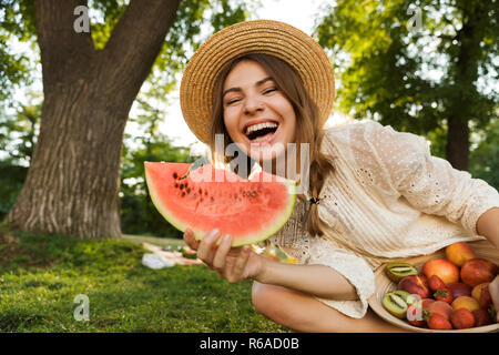 Smiling young girl in summer hat having a picnic at the park, sitting on a grass, eating watermelon and fruits from a bowl Stock Photo