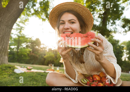 Happy young girl in summer hat having a picnic at the park, sitting on a grass, eating watermelon and fruits from a bowl Stock Photo