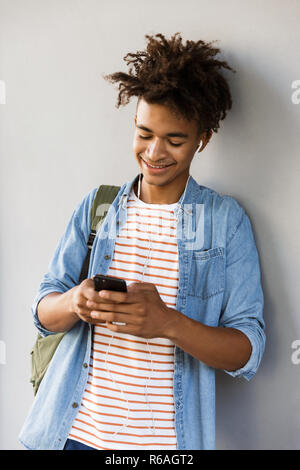 Smiling young african man with backpack outdoors, listening to music with earphones, using mobile phone Stock Photo