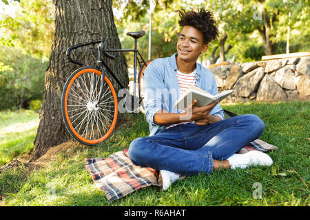 Smiling young african teenager with backpack outdoors, sitting on grass, holding book Stock Photo