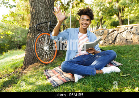 Positive young african teenager with backpack outdoors, sitting on grass, holding book Stock Photo