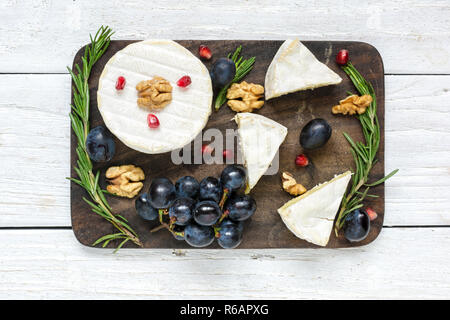 camembert cheese with grapes, pomegranate seeds, walnuts and rosemary on wooden cutting board. top view Stock Photo