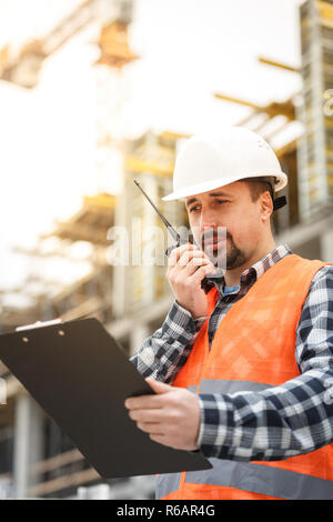 Developing engineer wearing white safety vest and hardhat with walkie talkie and clipboard inspecting construction site. Development and construction  Stock Photo