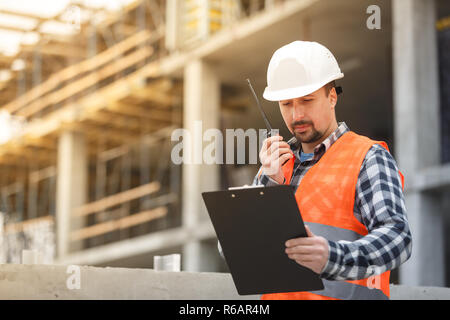 Developing engineer wearing white safety vest and hardhat with walkie talkie and clipboard inspecting construction site. Development and construction  Stock Photo