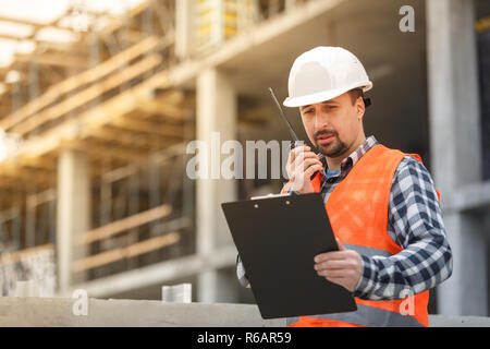 Developing engineer wearing white safety vest and hardhat with walkie talkie and clipboard inspecting construction site. Development and construction  Stock Photo