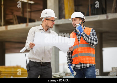 Male architect and developer with walkie talkie discussing blueprints of an architect project at  construction site Stock Photo