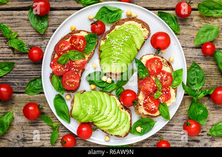 wholegrain bread avocado and tomatoes toasts with hummus, sesame and basil in a plate over rustic wooden background. healthy vegan food. top view Stock Photo
