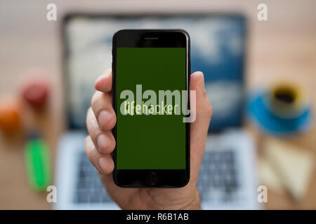 A man looks at his iPhone which displays the LifeHacker logo, while sat at his computer desk (Editorial use only). Stock Photo