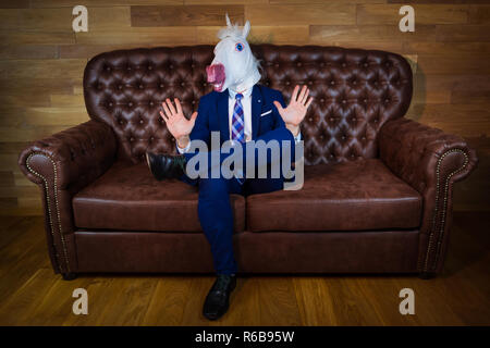 Funny unicorn in elegant suit siting on sofa like a boss and showing do not bother me gesture. Portrait of unusual man at home. Freaky guy in mask. Stock Photo