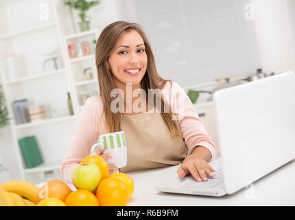Beautiful young woman in the kitchen drinking coffee and reading recipe on laptop. Looking at camera. Stock Photo