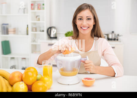 Beautiful young woman in the kitchen squeezing orange juice with an electric juicer. Looking at camera. Stock Photo