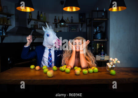 Funny couple playing at bar counter in kitchen with apples on table. Blonde girl have fun with boyfriend in mask. Unicorn in suit with young woman Stock Photo