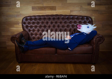 Funny unicorn in elegant suit lies on leather sofa. Unusual man relaxing at home. Freaky guy in comical mask on background of wooden wall. Stock Photo