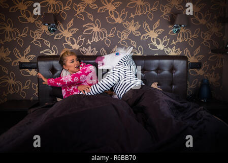 Angry man in comical mask molest with scared young woman in pajama on the bed. Unusual couple has relationship problems in stylish bedroom. Stock Photo