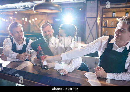 Group of positive happy waiters in uniform leaning on bar counter and clinking beer bottles while celebrating success after hours in restaurant Stock Photo
