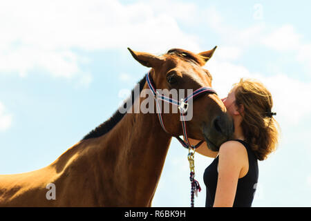 Taking care of animals, love and friendship concept. Jockey young girl kissing and hugging brown horse on sunny day Stock Photo