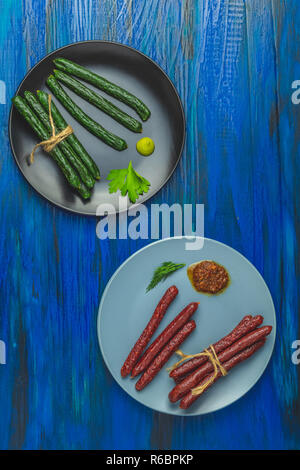 Kabanosy, sausages green with wasabi made of pork in black plate and traditional polish in gray plate on a dark blue wood surface with addition of fre Stock Photo
