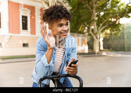 Smiling young african man outdoors, riding on a bicycle, waving, using mobile phone Stock Photo