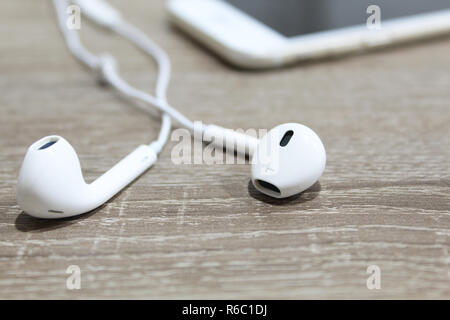 White earphones on wooden surface and smartphone in background. Small earbuds close up Stock Photo