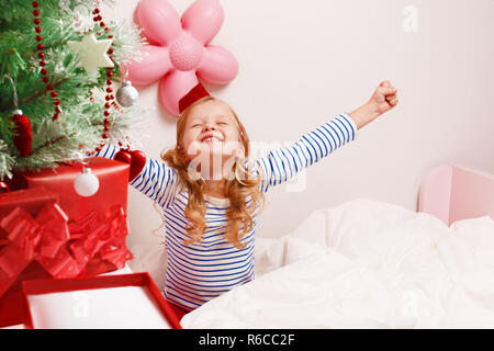 Happy little child girl in santa hat pulls hands up sitting in bed after waking up. Christmas morning. Stock Photo
