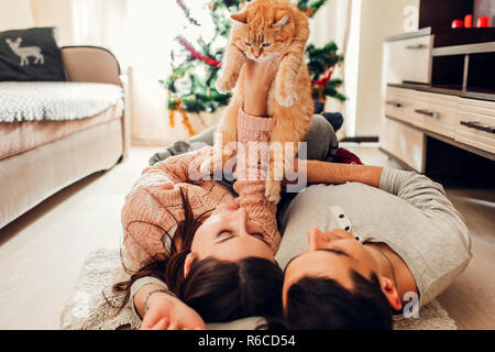 Couple in love lying by Christmas tree and playing with cat at home. Man and woman lifting pet in hands. Happy family Stock Photo