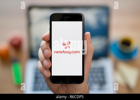 A man looks at his iPhone which displays the Nearbuy logo, while sat at his computer desk (Editorial use only). Stock Photo