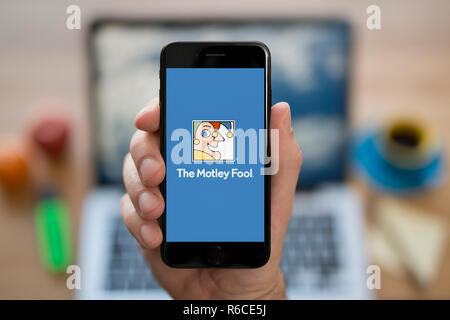 A man looks at his iPhone which displays The Motley Fool logo, while sat at his computer desk (Editorial use only). Stock Photo