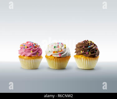 Three cupcakes with frosting and colourful sweets on a plain surface Stock Photo