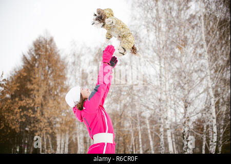 dog throws up into the sky. Walk in the winter outdoors with a dog breed Shih Tzu. A woman in a bright red warm ski clothing walking in the snow with your pet, a little shih tzu dressed in overalls. care for animals, and loves playing with the dog Stock Photo