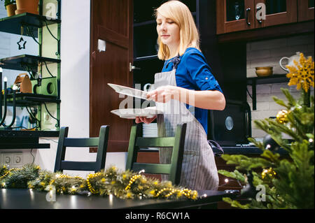 woman housewife setting the table. young blonde in a dress and apron, served in a dining table, puts the dishes, doing the work in the kitchen Stock Photo