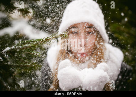 Blowing Snow. Beautiful Joyful Female Model on background of fir trees. Young woman enjoying magical snowfall. Winter wonderland. Stock Photo