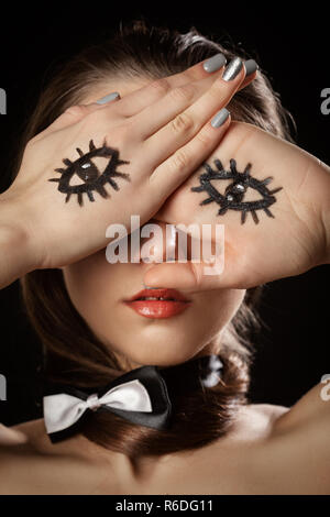 young woman with tie butterfly cover face with palms with drawn eyes, black background Stock Photo