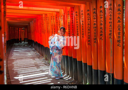 Woman Dressed In Traditional Japanese Costume Walking Under Tori Gates At The Fushimi-Inari Shrine, Kyoto Japan Stock Photo