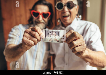 Two elderly male friends with party sunglasses showing their picture. Senior people having great time together. Focus on picture in hand of old people Stock Photo