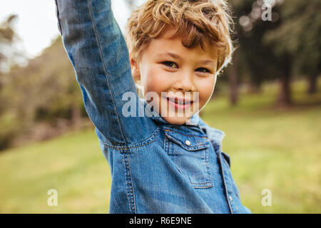 Close up of cute little boy standing outdoors with his hand raised. Small kid wearing a denim shirt looking at camera. Stock Photo